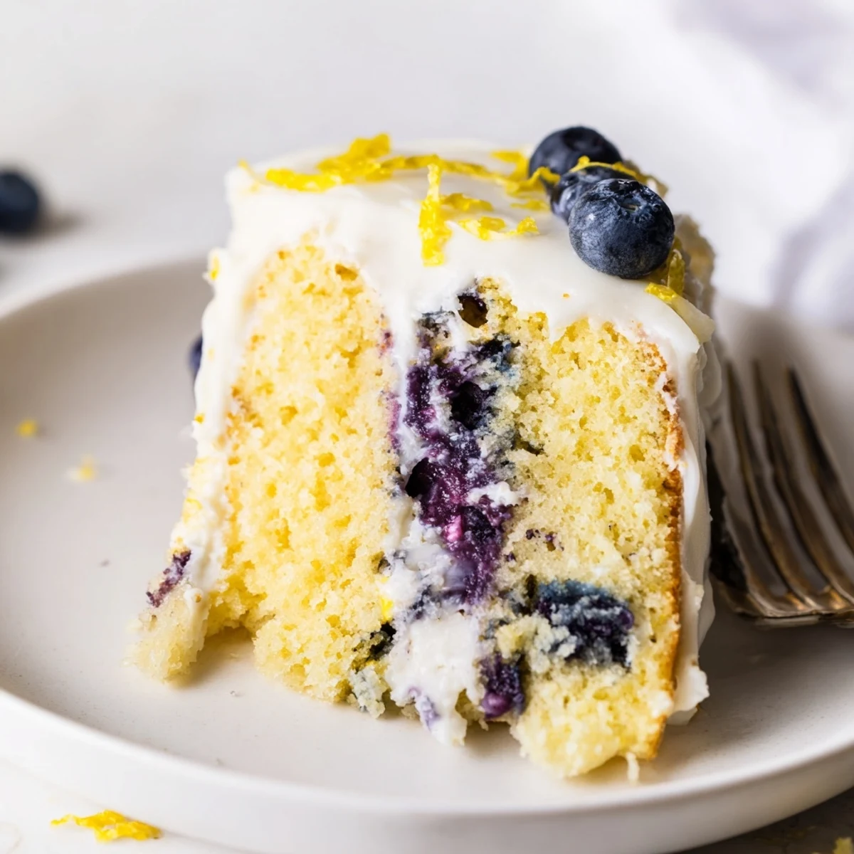 A whole Lemon Blueberry Cake with Frosting sits on a cake stand, topped with fresh blueberries and lemon zest for a spring dessert.