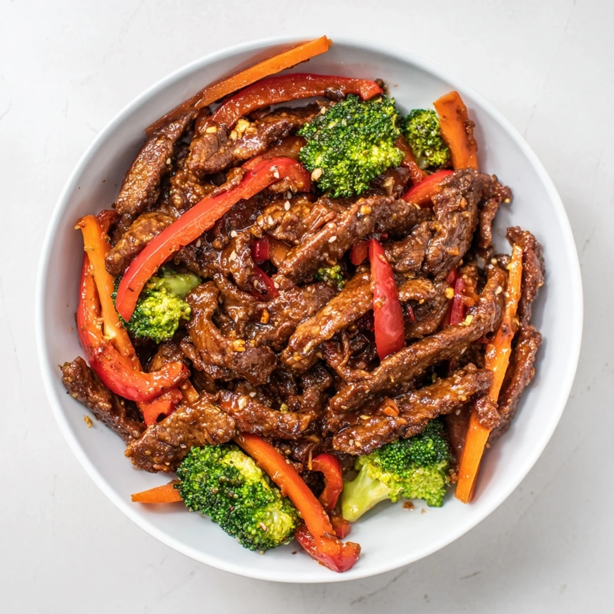 Tender beef strips and crisp veggies in a spicy stir fry sauce glisten in a white ceramic bowl.