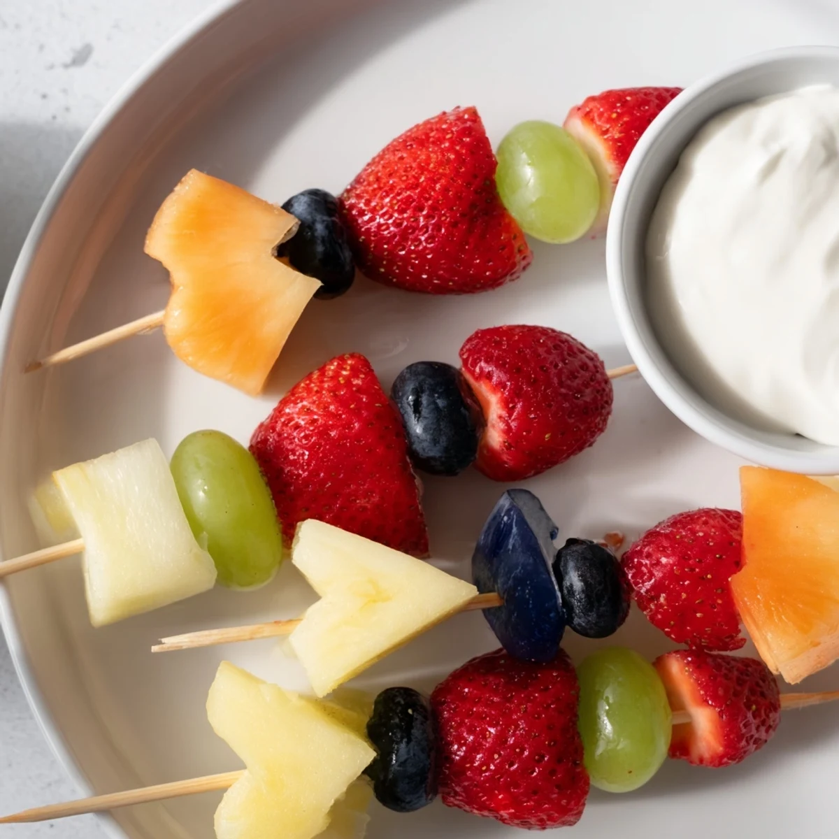 Bright Sweetheart Fruit Kabobs with Yogurt are displayed on a white plate, with Greek yogurt dip topped with a blueberry garnish.