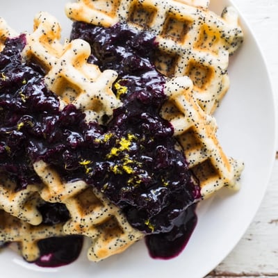 A close-up of fluffy waffles with lemon zest and poppy seeds, drizzled in thick, homemade blueberry syrup.  
